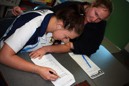 Female student writing at desk with another student sitting with her.