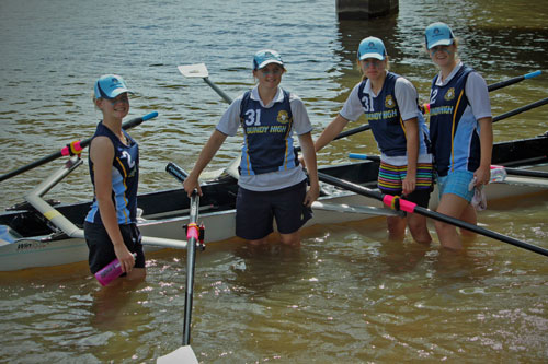 Four female students in sport uniform standing in water next to a row boat.